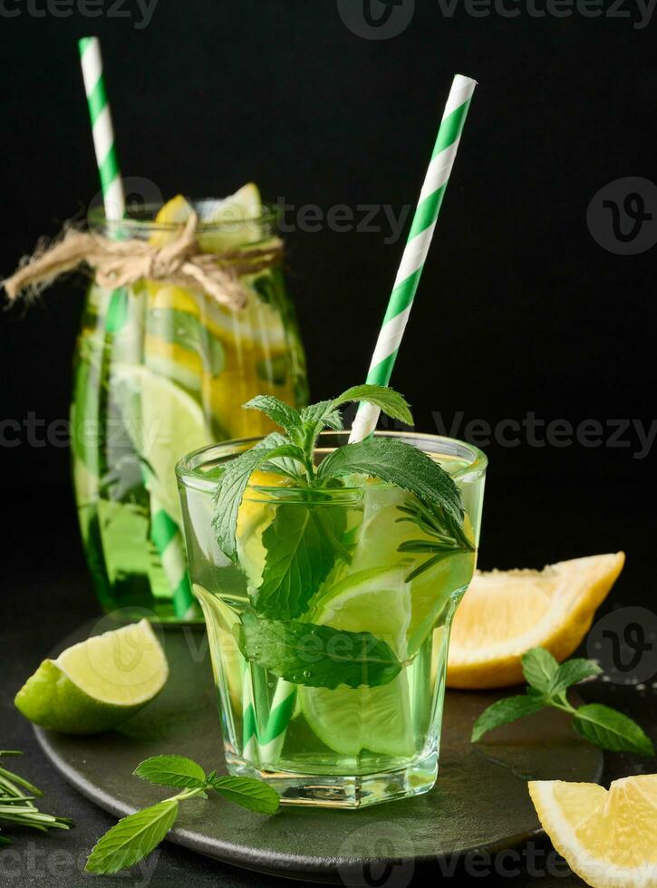 Lemonade in a transparent glass with lemon, lime, rosemary sprigs and mint leaves on a black background photo