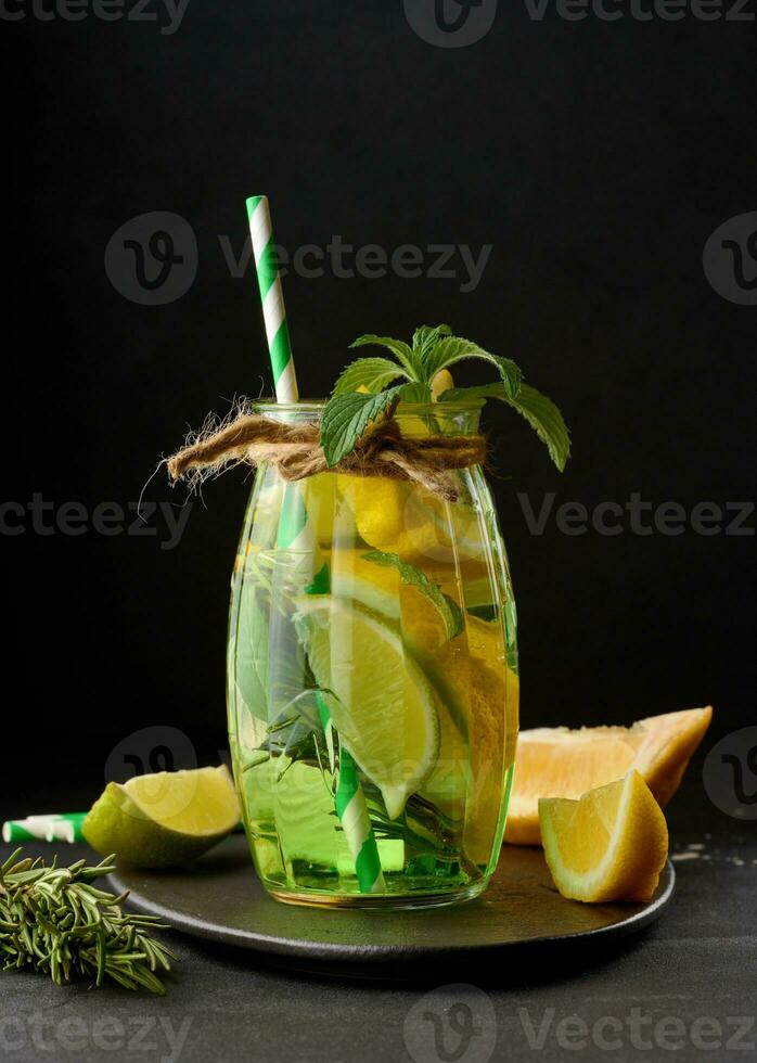 Lemonade in a transparent glass with lemon, lime, rosemary sprigs and mint leaves on a black background photo