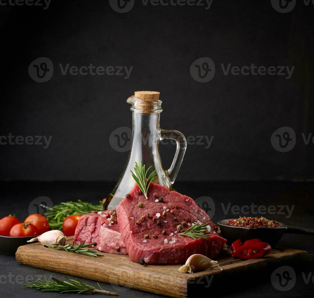 Raw piece of beef with spices pepper, rosemary sprig, salt and olive oil on a wooden board, black background photo