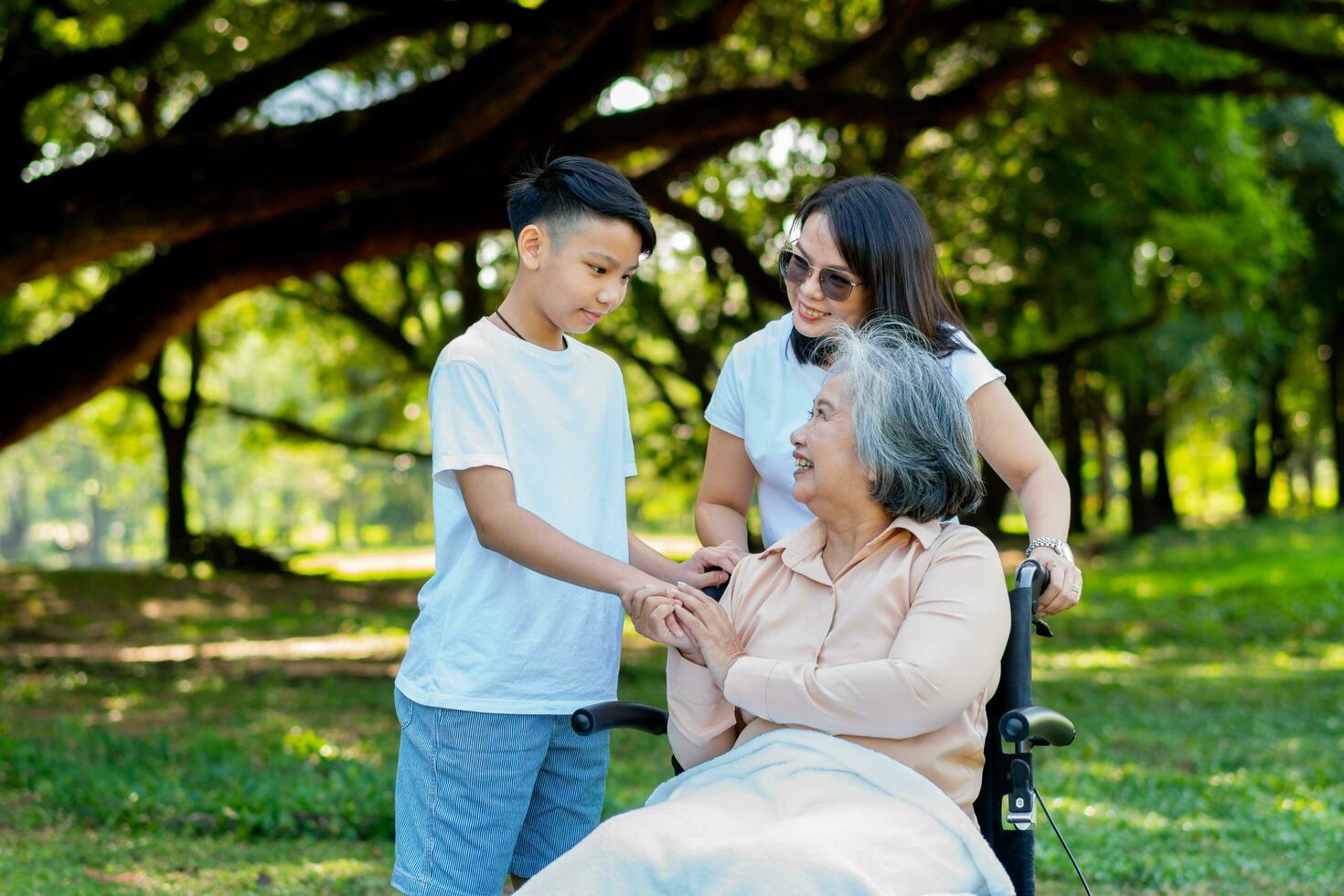 Happy senior Asian grandmother uses wheelchair with her daughter and grandchild in park, Grandson came to visit elderly grandmother and hold hand. Concept of happy family, good relationship together photo