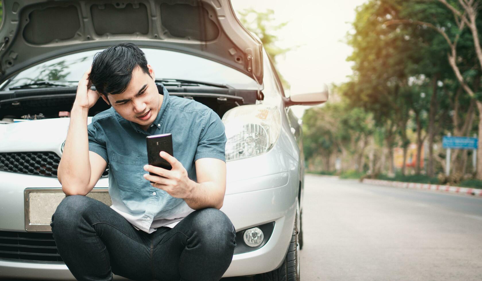 Asian man using mobile phone calling for assistance after a car breakdown on street. Concept of vehicle engine problem or accident and emergency help from Professional mechanic photo