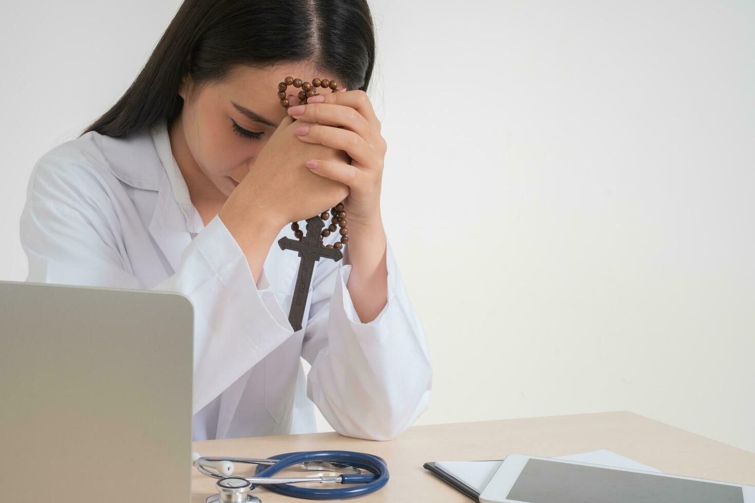 Young Asian woman doctor sitting on desk and holding a cross and praying, she looks unhappy and tired because of overwork, Concept of stressed exhausted and doctor liability, Dealing with frustrated photo