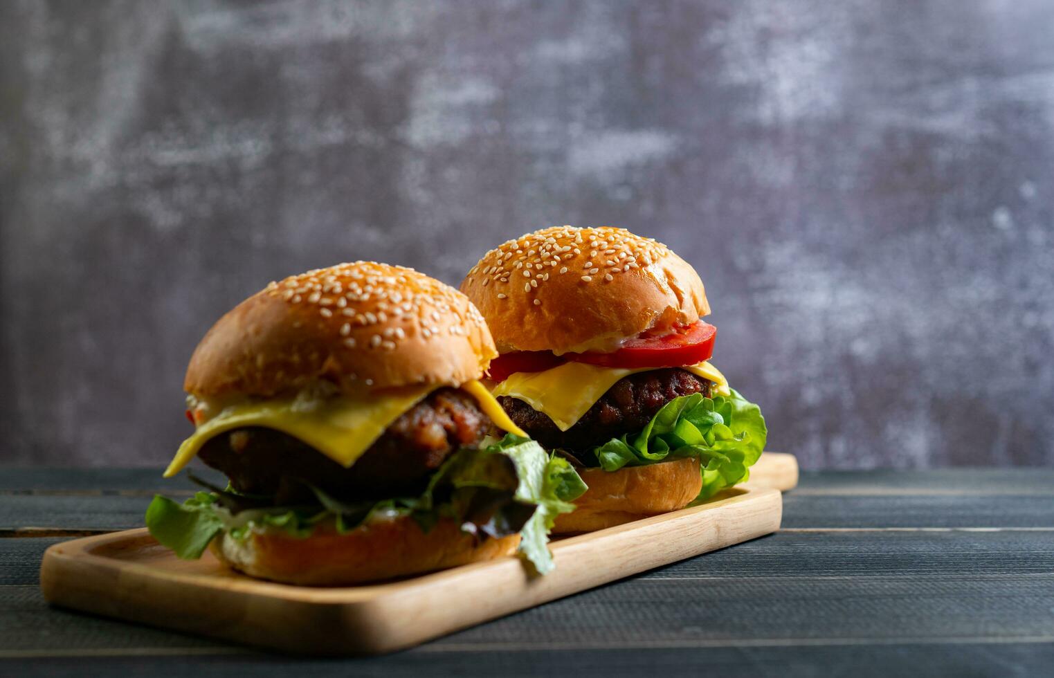 Two fresh tasty homemade hamburgers with fresh vegetables, lettuce, tomato, cheese beside sliced tomatoes on a cutting board. Free space for text photo