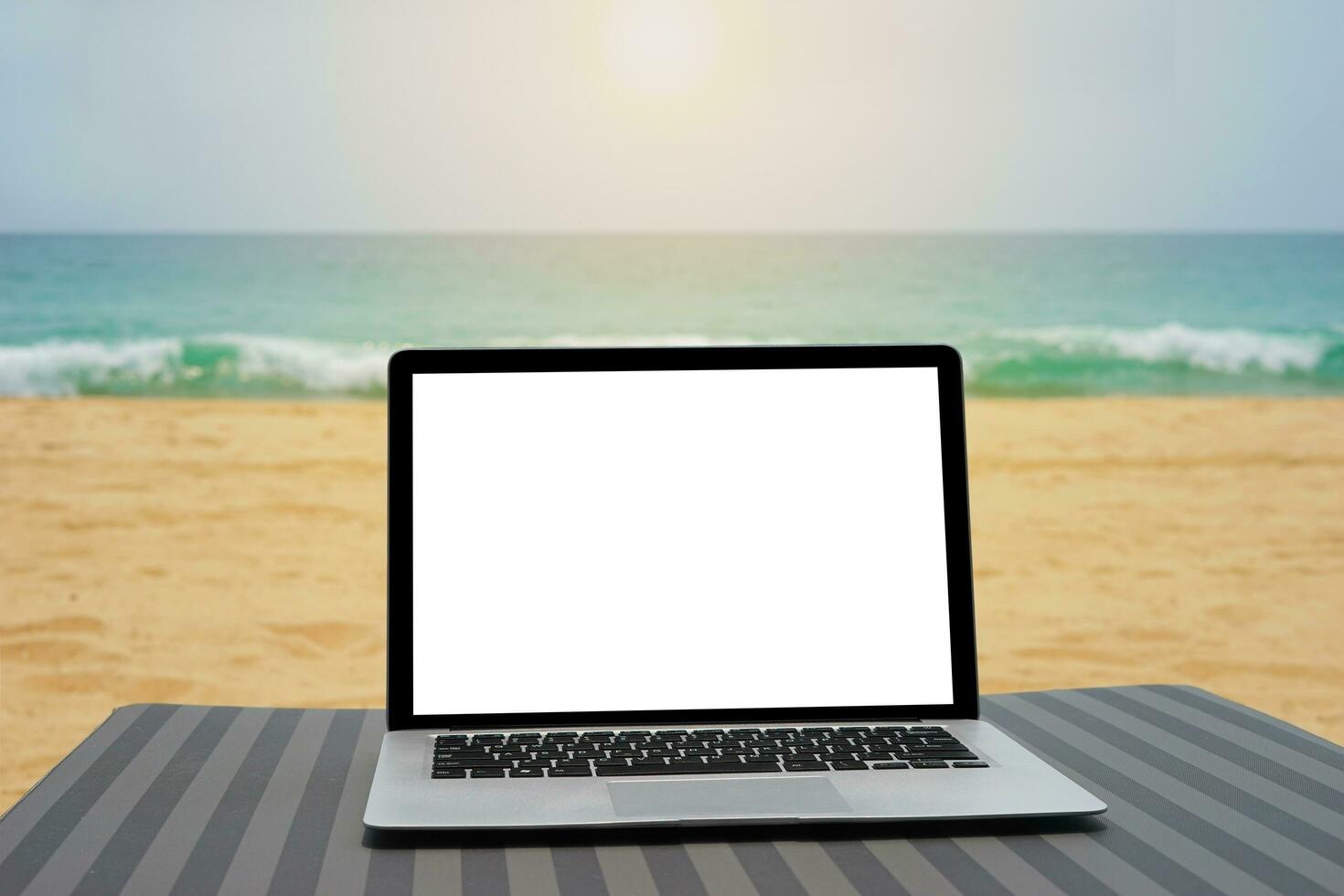 Laptop with blank screen for creative design on beach bed nearby beach and blue sea background. Computer notebook with clipping path for present landing page design mock up template photo