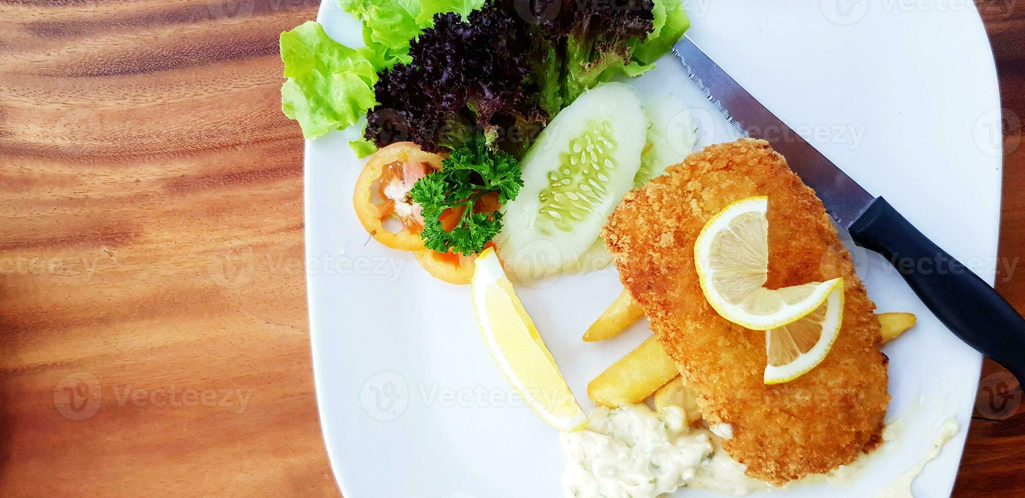 Fried Dolly Fish Steak with fresh organic vegetable, green cabbage, sliced cucumber, tomato and lemon, french fried, cream sauce and knife in white plate on wooden table with left copy space. photo