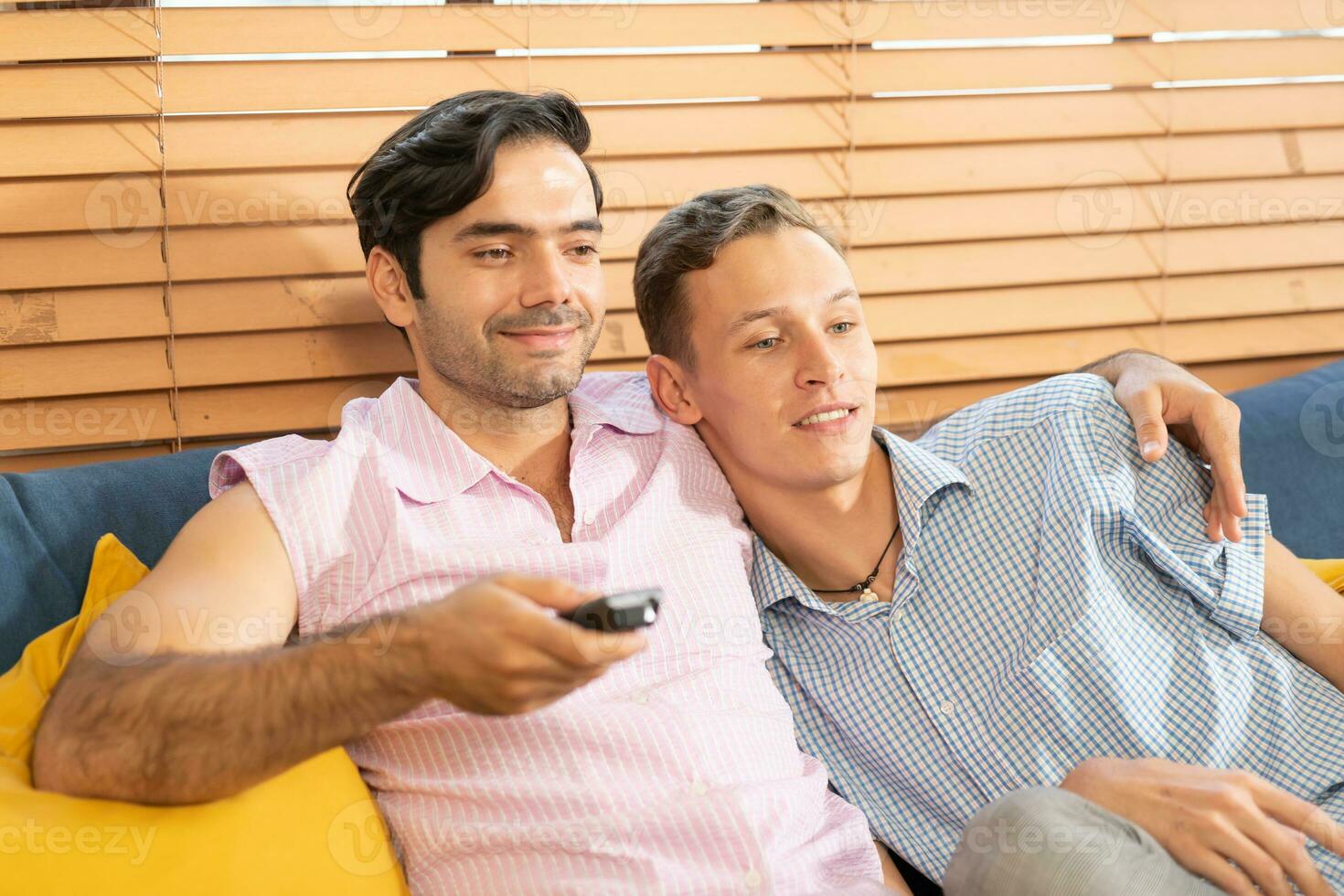 dos hombres en casual trajes sentado en sofá abrazando y acecho televisión con cerveza y Pizza en vivo habitación a hogar. lgbt gay relación concepto. foto