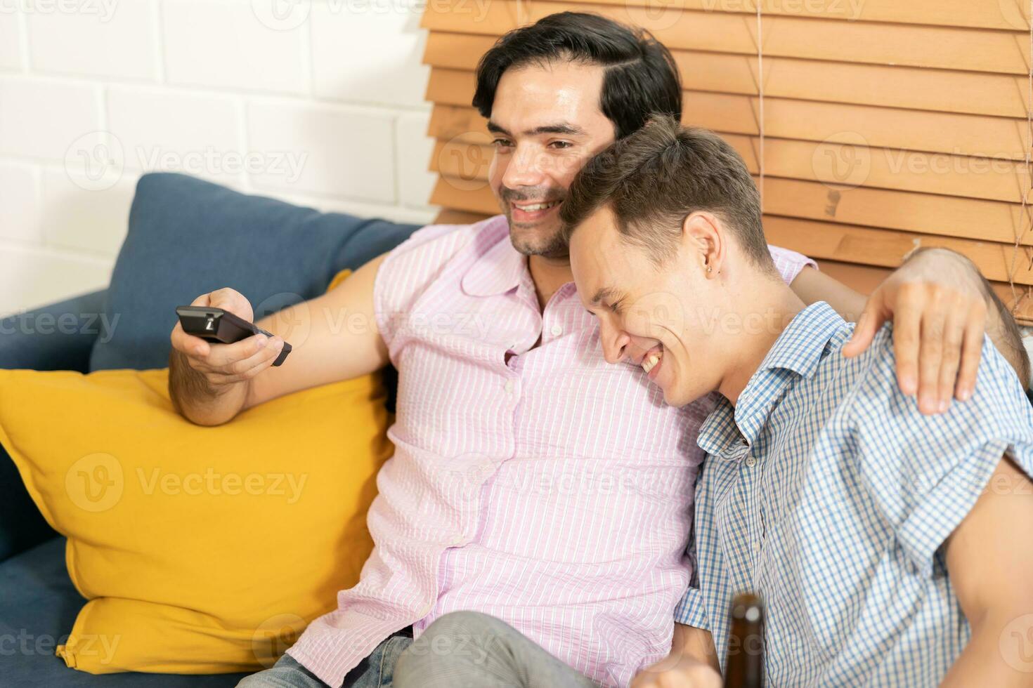 dos hombres en casual trajes sentado en sofá abrazando y acecho televisión con cerveza y Pizza en vivo habitación a hogar. lgbt gay relación concepto. foto