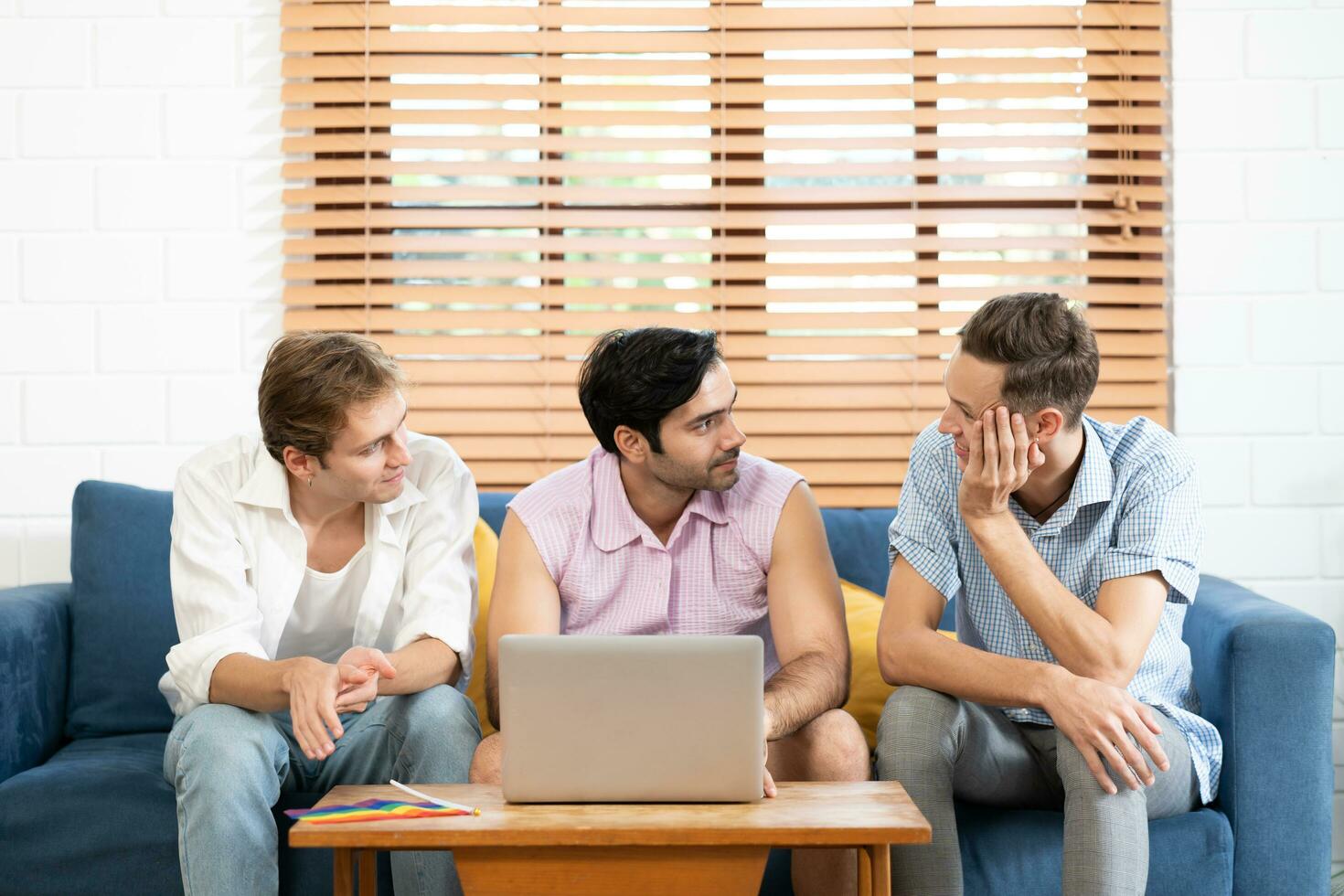 Triple young man sitting in the living room while using laptop to video call with rainbow flag on desk. LGBTQ people lifestyle and love emotion. LGBT social network. selective focus photo