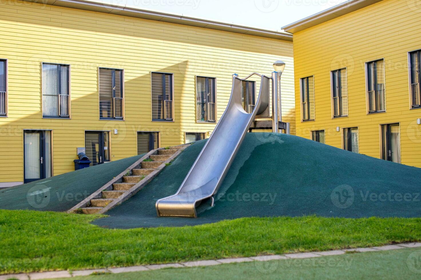 un metal diapositiva en un para niños patio de recreo con un verde césped. escalera a el colina. amarillo casa foto