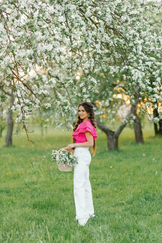 un hermosa joven europeo mujer con trenzas en el primavera en el parque sostiene un cesta de flores en un floreciente huerta de manzana arboles foto