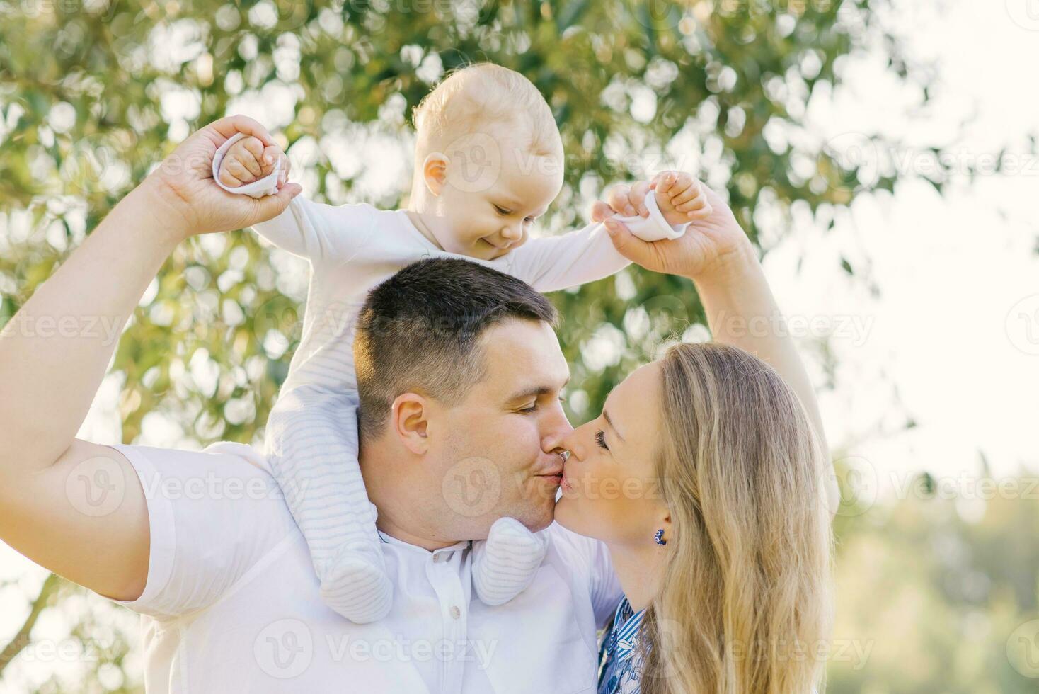 A European family in the park on a summer day. Mom, dad and little son in their arms. Parents kiss each other photo