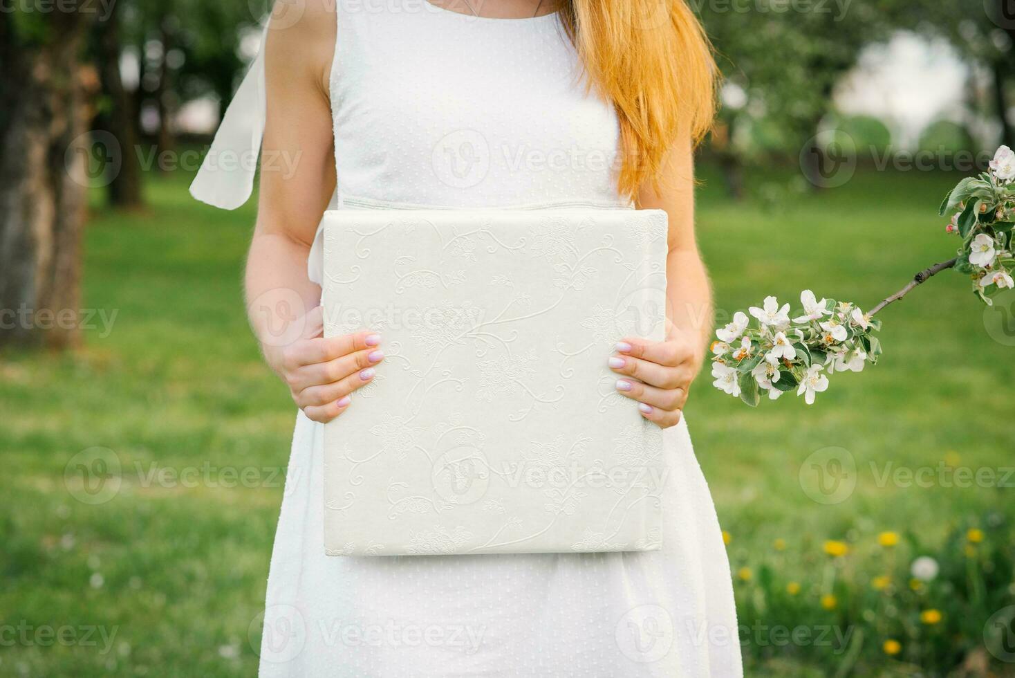 A photo book in a white leather cover in the hands of a woman in spring near a flowering branch of an apple tree