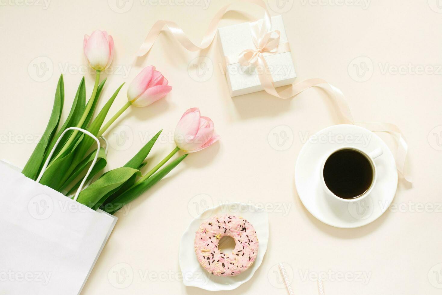 Morning cup of coffee, donut, gift or present box and spring tulip flowers on beige background. Beautiful breakfast for Women day, Mother day, Valentine's Day. Flat lay. photo