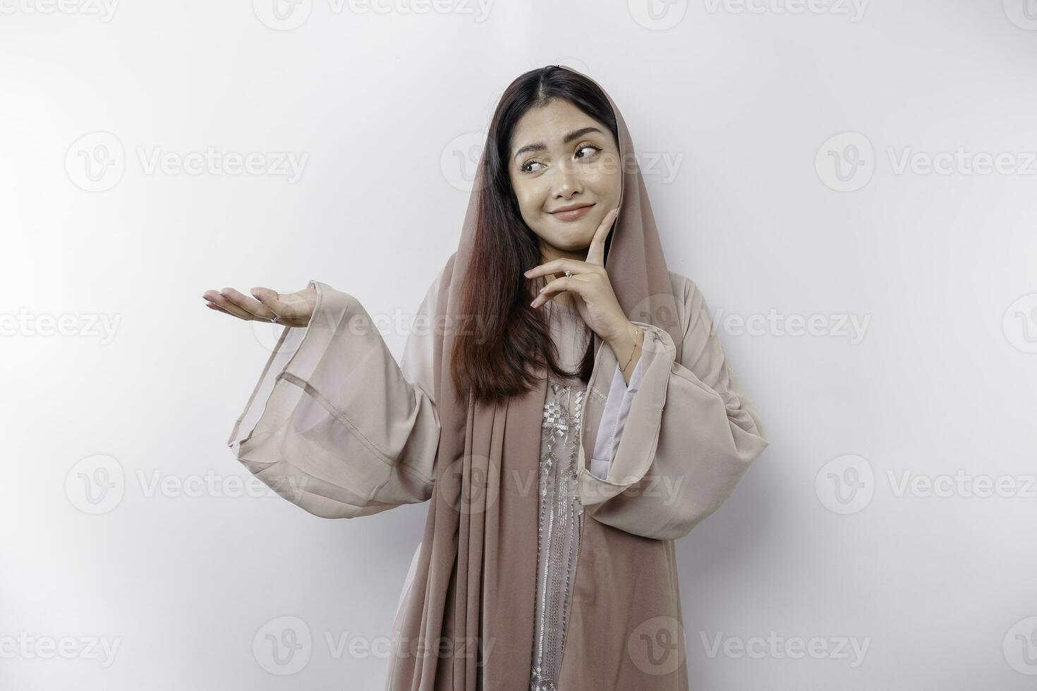 A thoughtful young Asian Muslim woman is wearing hijab and looks confused between choices, isolated by a white background photo