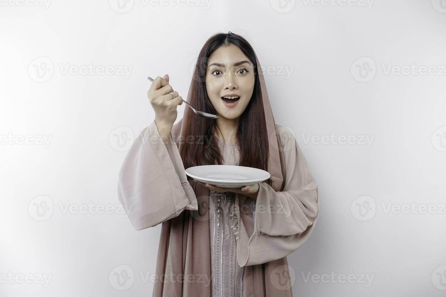 A smiling Asian Muslim woman is fasting and hungry and holding and pointing to a plate photo