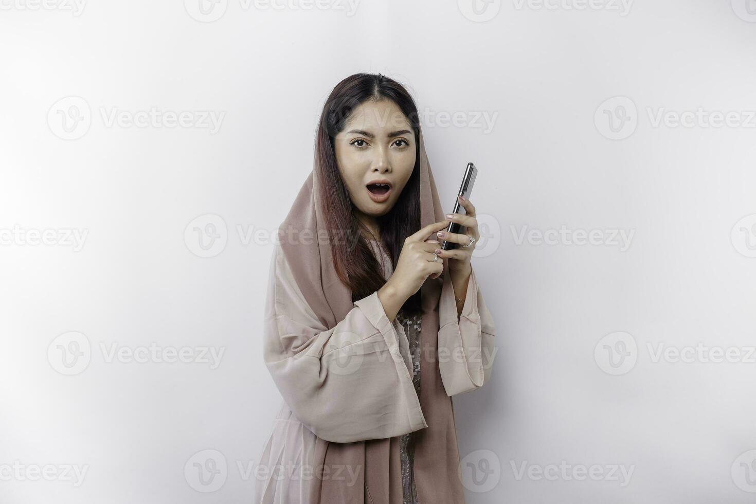 Shocked Asian woman wearing headscarf, holding her phone, isolated by white background photo