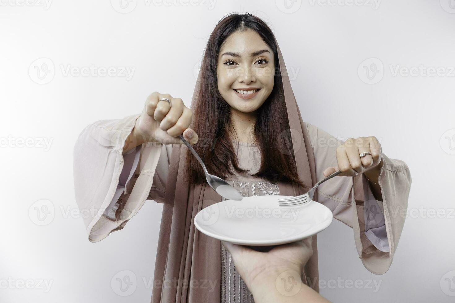 A smiling Asian Muslim woman is fasting and hungry and holding and pointing to a plate photo