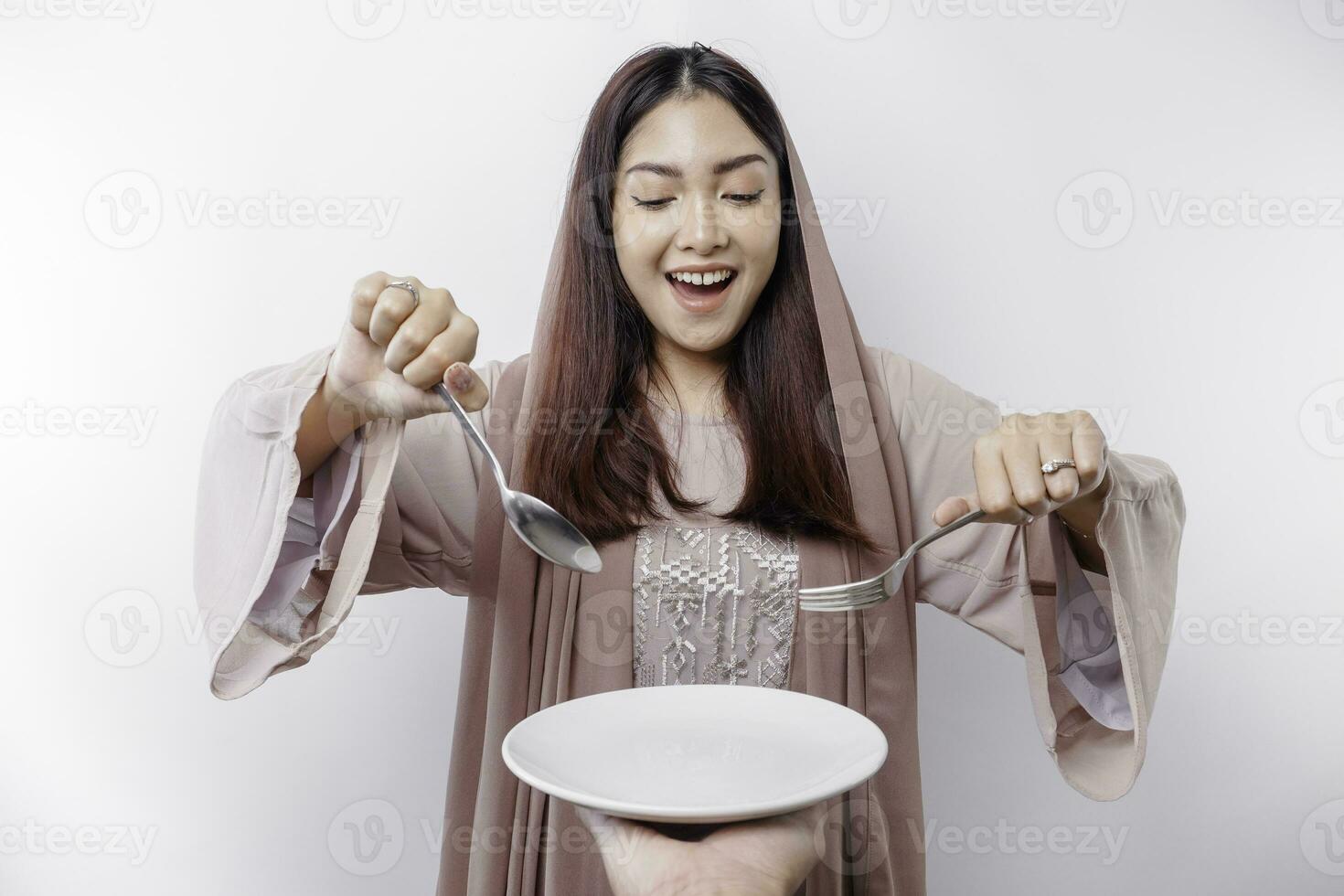 A smiling Asian Muslim woman is fasting and hungry and holding and pointing to a plate photo