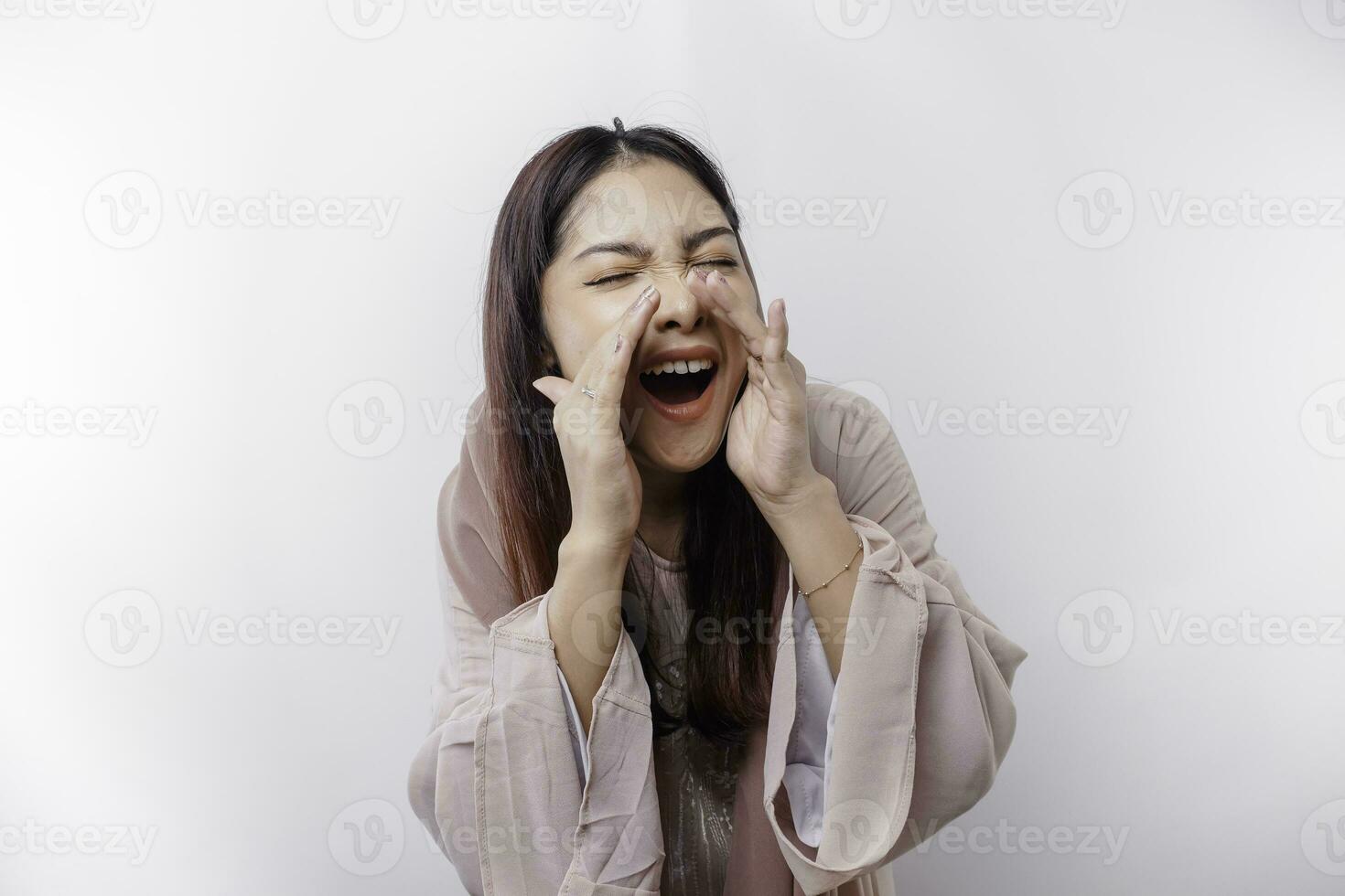 Young beautiful Asian Muslim woman wearing a headscarf shouting and screaming loud with a hand on her mouth. communication concept. photo