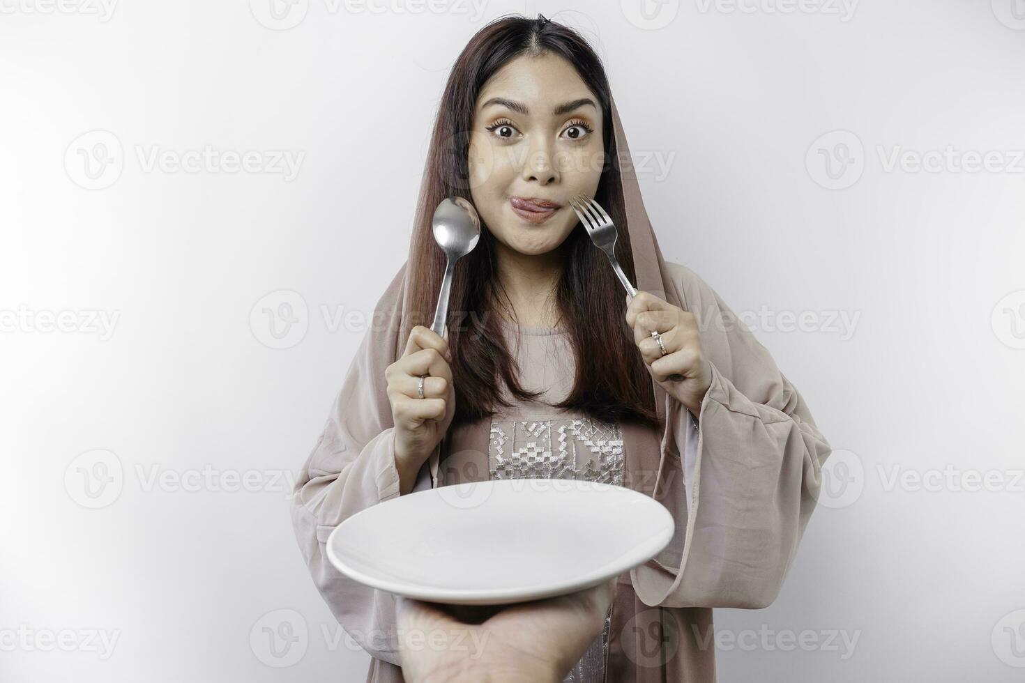 A smiling Asian Muslim woman is fasting and hungry and holding and pointing to a plate photo