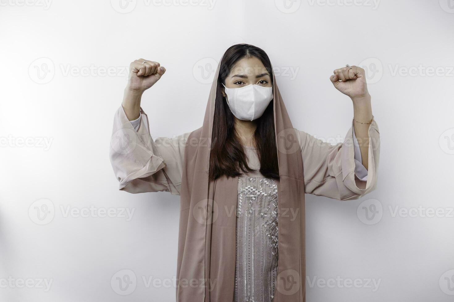 An Asian Muslim woman wearing a mask as a preventive for covid-19 is expressing a happy successful expression wearing a hijab isolated by white background photo