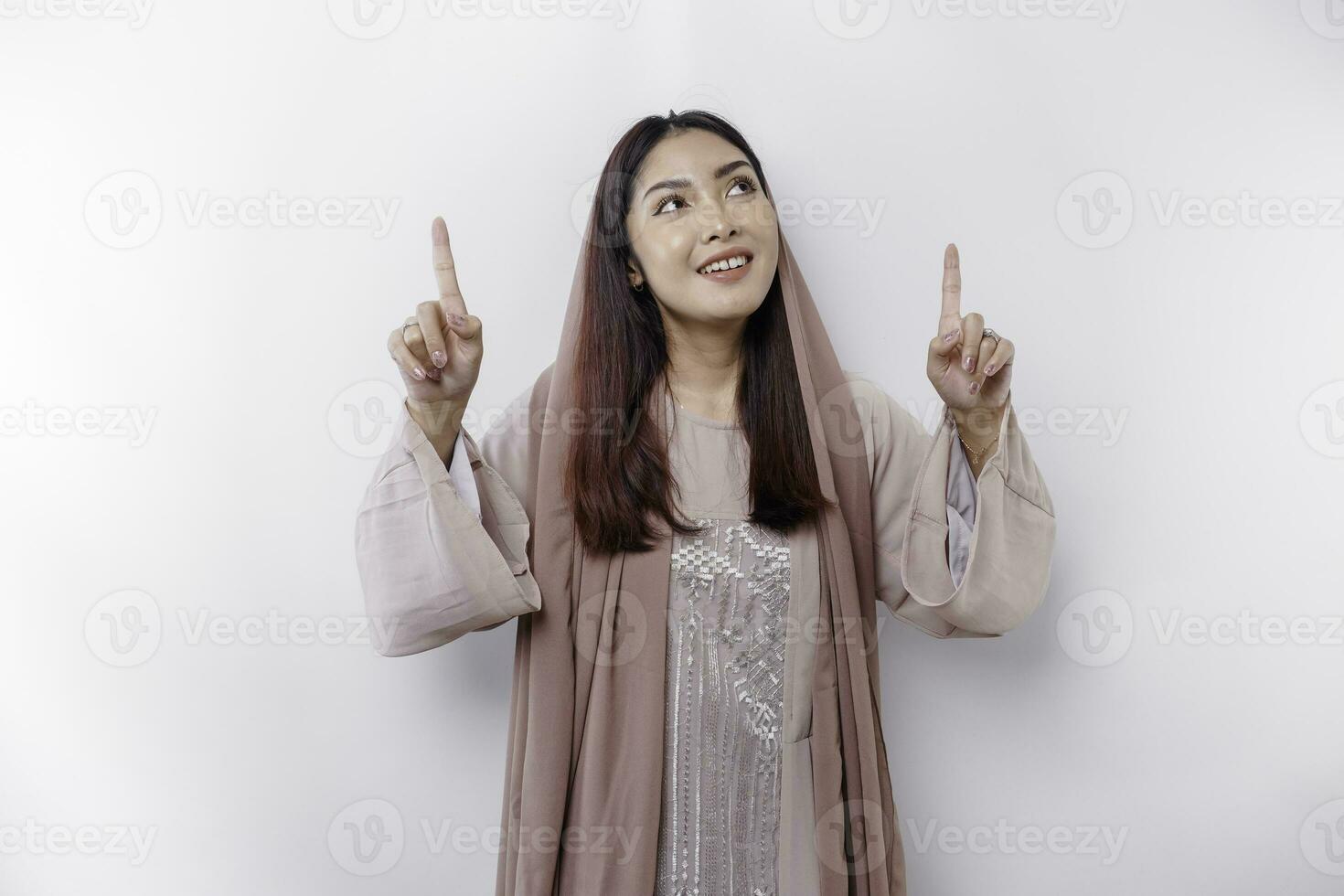 Excited Asian Muslim woman wearing headscarf pointing at the copy space above her, isolated by white background photo