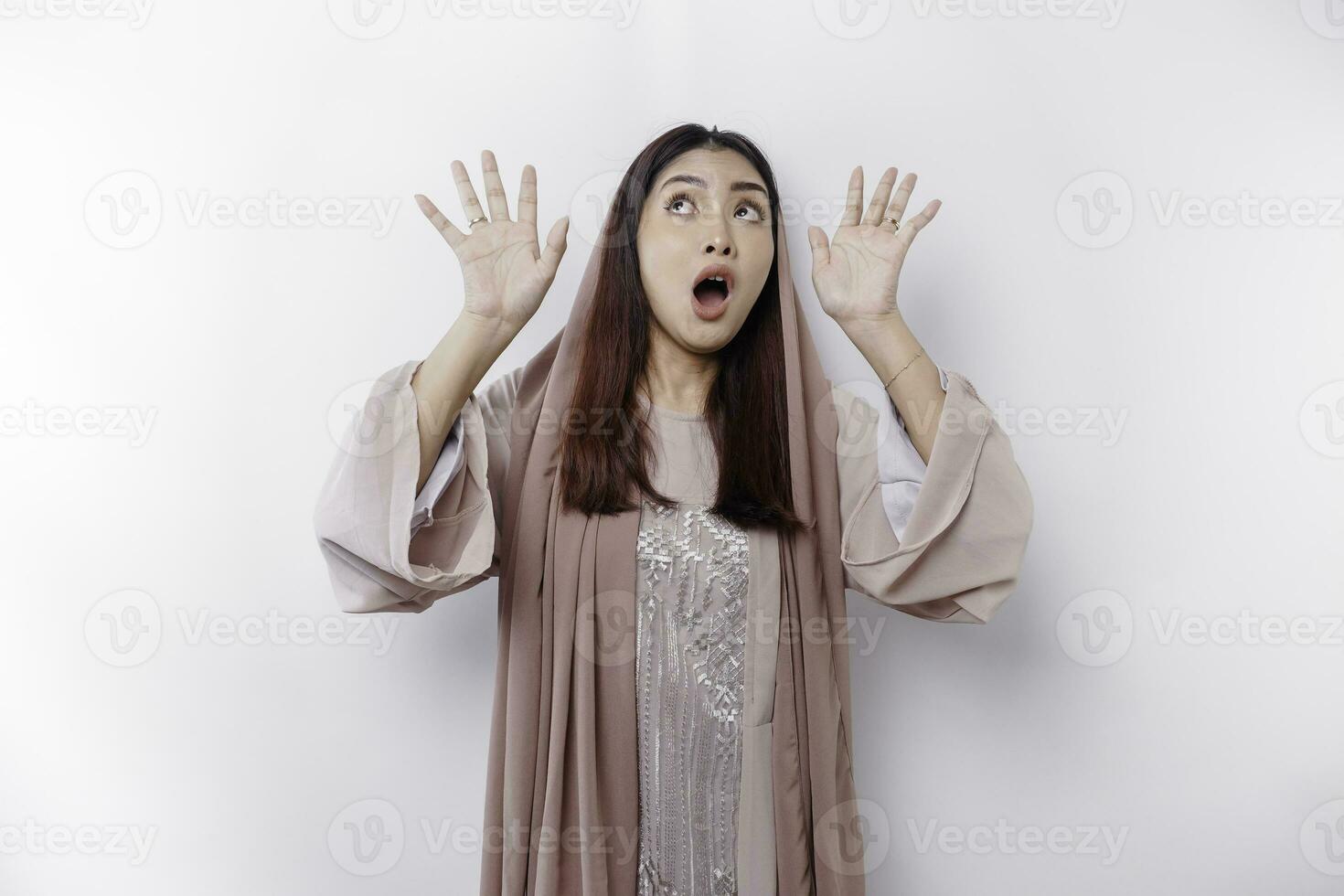 Shocked Asian Muslim woman wearing a headscarf pointing at the copy space on top of her, isolated by a white background photo