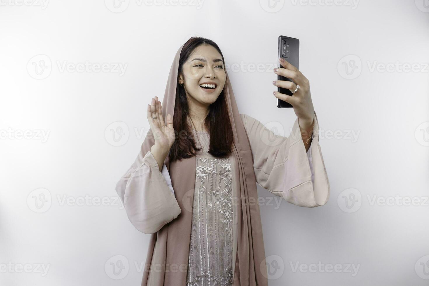 A happy Asian Muslim woman wearing a headscarf, happily talking during video call, isolated by white background photo