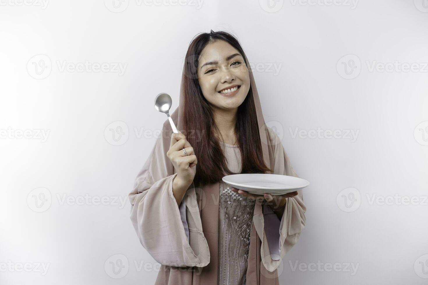 A smiling Asian Muslim woman is fasting and hungry and holding and pointing to a plate photo