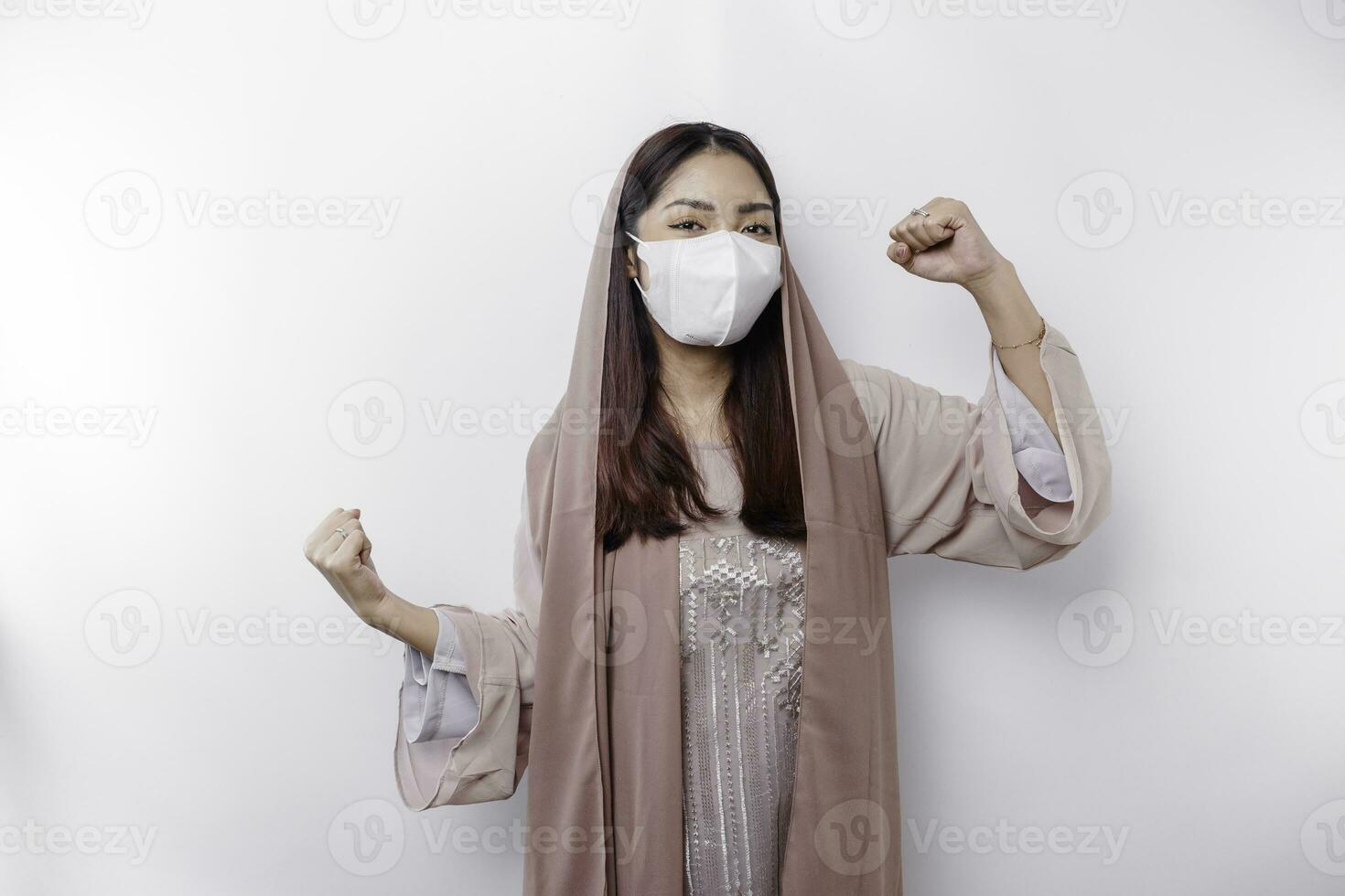 An Asian Muslim woman wearing a mask as a preventive for covid-19 is expressing a happy successful expression wearing a hijab isolated by white background photo