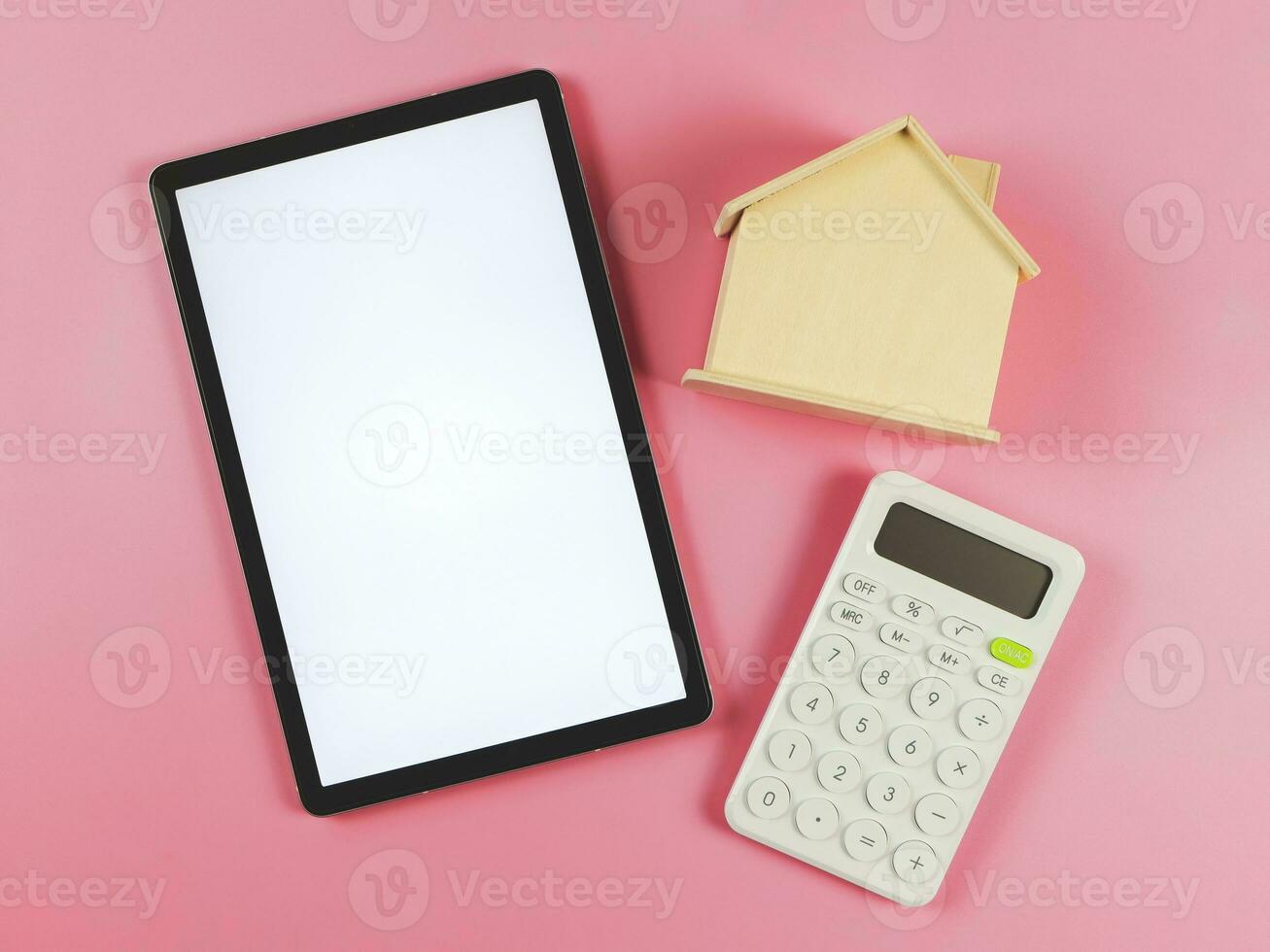 flat layout of digital tablet with white blank screen, wooden house model  and white calculator on pink  background with copy space, home purchase concept. photo