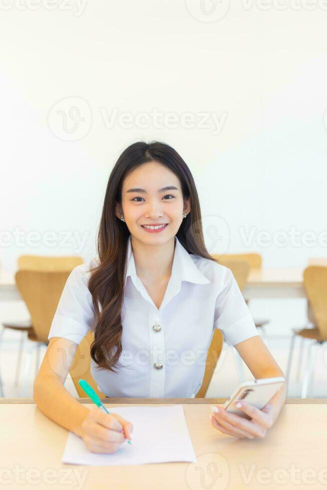 joven asiático mujer estudiante en uniforme utilizando teléfono inteligente y escritura alguna cosa acerca de Trabaja ahí son muchos documentos en el mesa su cara con sonriente en trabajando a a buscar información para estudiar foto