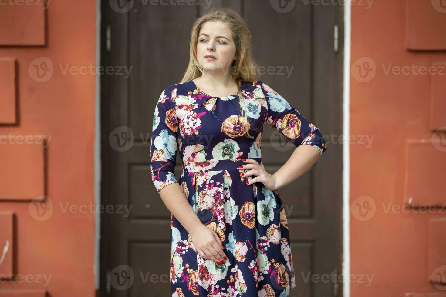 A blonde girl in a blue floral dress stands near a brown door. photo