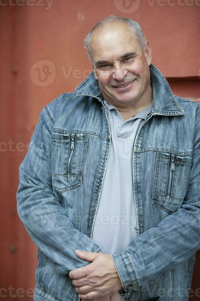 An elderly man in a denim jacket looks at the camera and smiles. An upright fifty-year-old gray-haired man of large build. photo