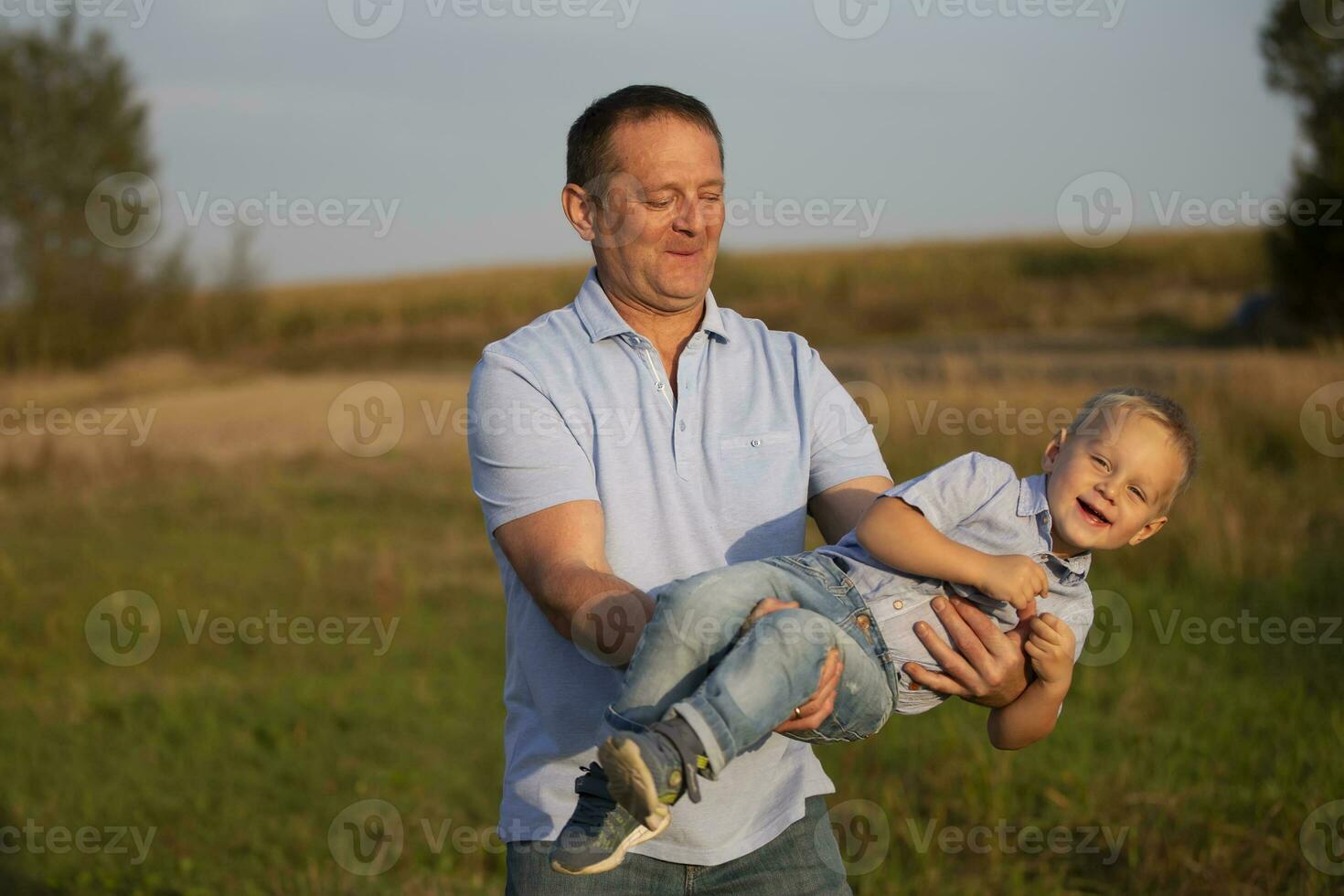Happy dad plays with his son on the background of the field. The joyous grandfather twirls his grandson in his arms. photo