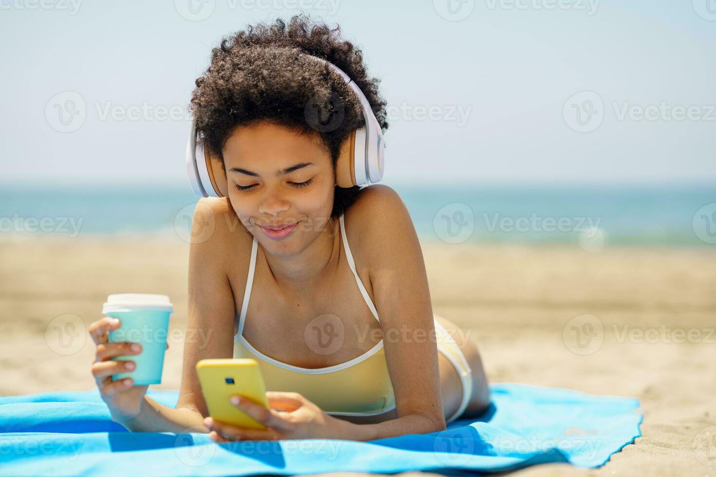 Happy black lady using smartphone and drinking takeaway beverage lying on seashore photo