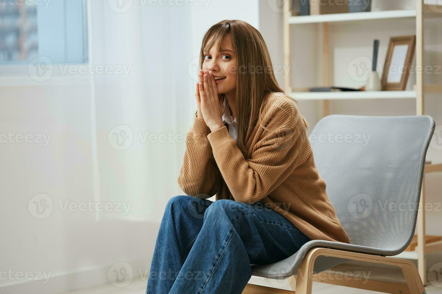 alegre linda joven rubia estudiante dama en calentar suéter doblez manos cerca cara sentado en Sillón a moderno hogar interior. pausa desde trabajar, tomar un romper, reflexiones en gratis hora concepto. Copiar espacio foto