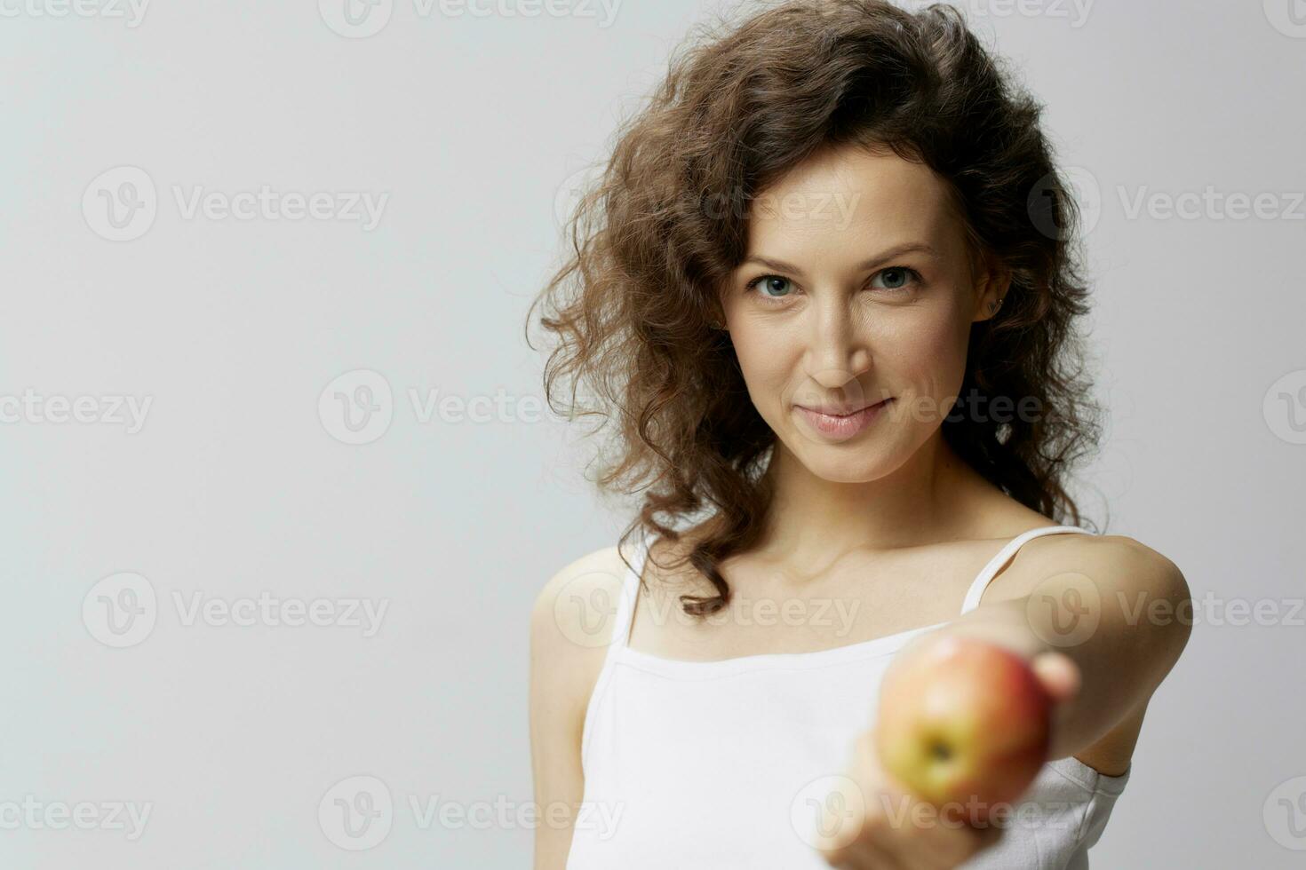 Smiling lovely cute curly beautiful woman in basic white t-shirt pulls apple enjoy healthy food posing isolated on over white background. Natural Eco-friendly products concept. Copy space photo