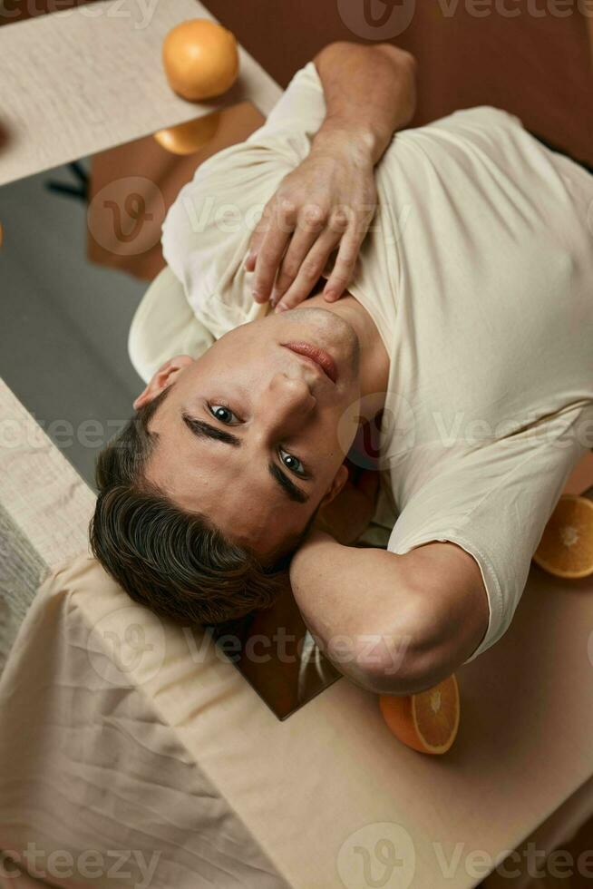 a young guy in a white jacket lies on a table with a mirror oranges photo