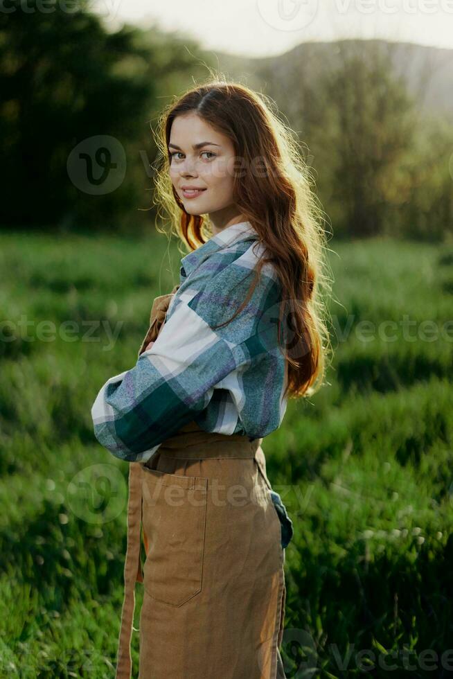 A woman gardener in an apron stands in a field of green grass outdoors, smiling on a summer day into a sunset photo