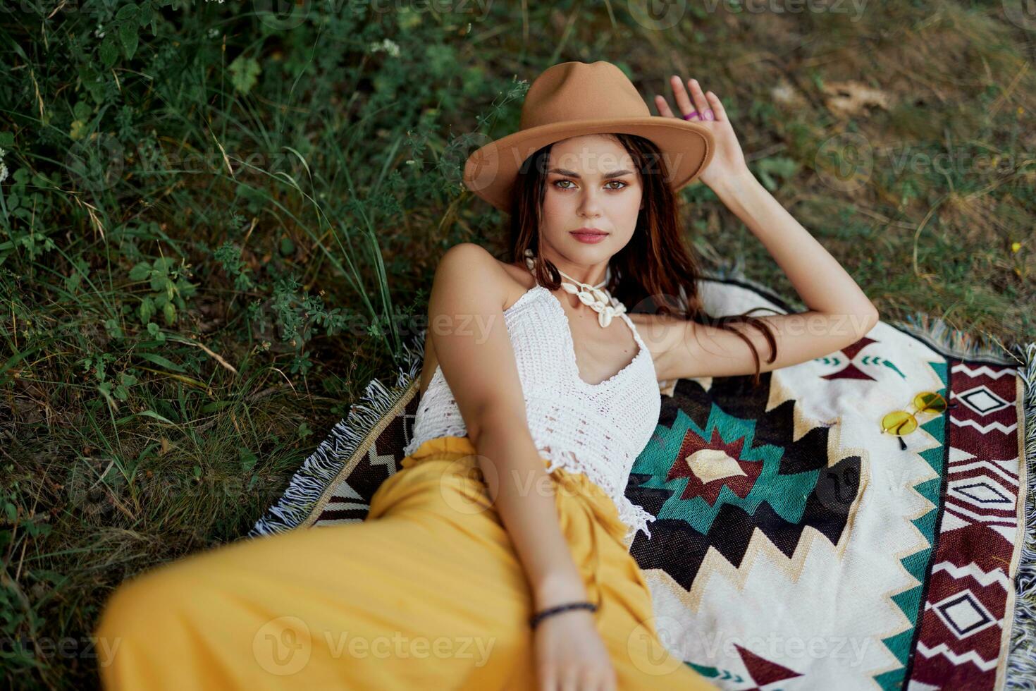 A woman in eco-friendly clothing in a hippie look is lying on a colored plaid smiling and looking at an autumn sunset in nature in the park. Lifestyle on a journey of harmony with the world photo