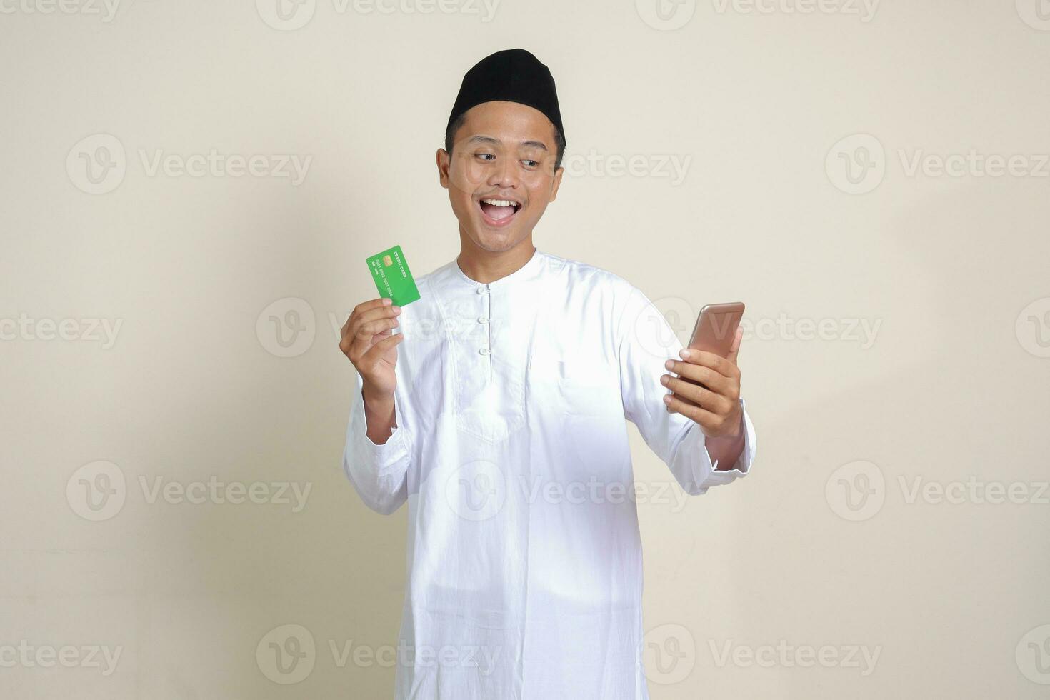 Portrait of attractive Asian muslim man in white shirt with skullcap holding a mobile phone and presenting credit card. Isolated image on gray background photo