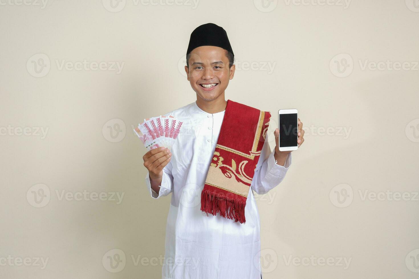 Portrait of attractive Asian muslim man in white shirt with skullcap showing one hundred thousand rupiah while showing blank screen mobile phone. Financial and shopping concept. Isolated image on gray photo