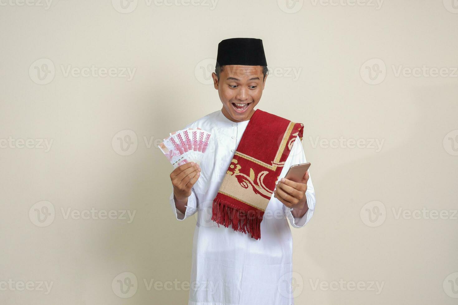 Portrait of attractive Asian muslim man in white shirt showing one hundred thousand rupiah while using mobile phone. Financial and savings concept. Isolated image on gray background photo