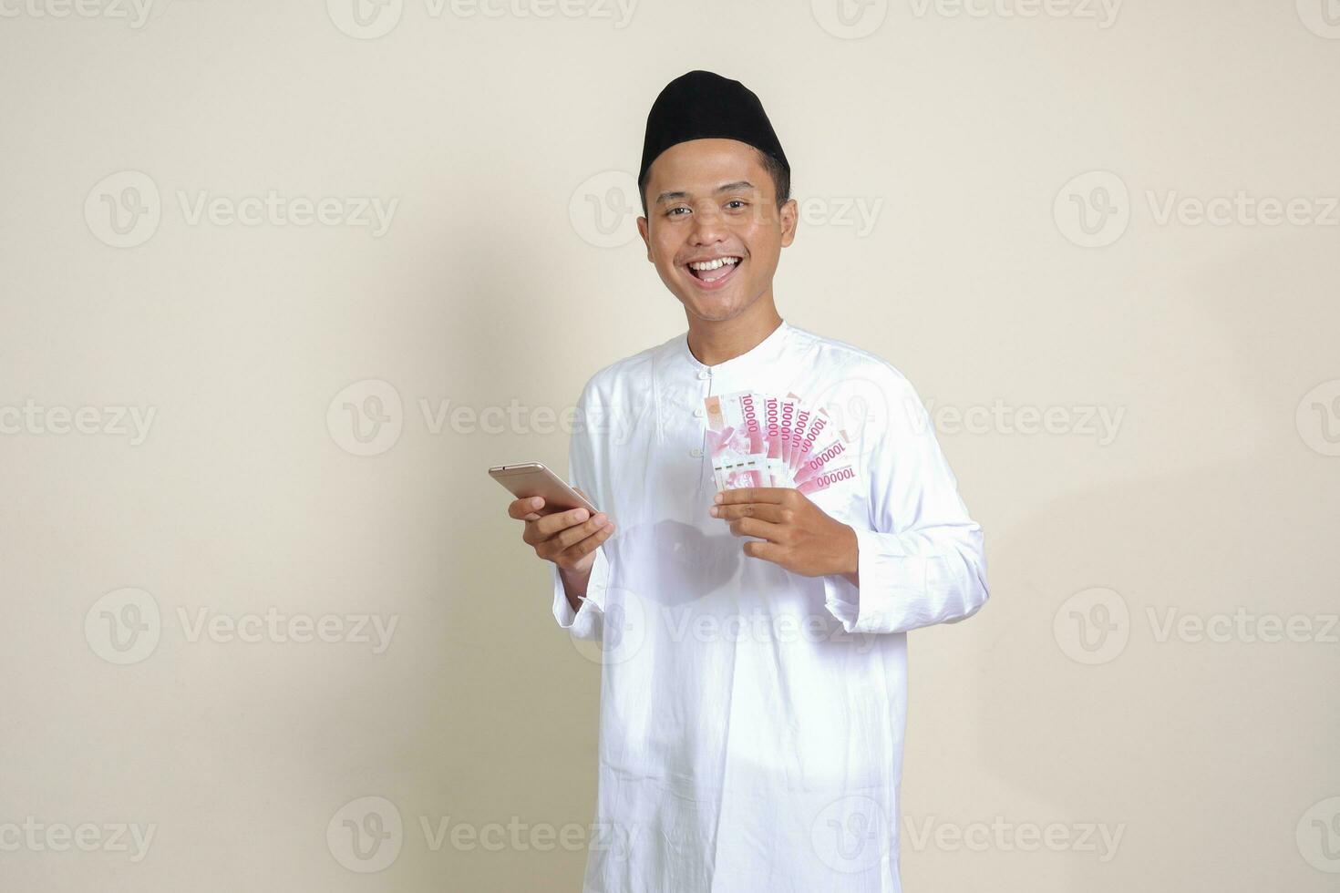 Portrait of attractive Asian muslim man in white shirt showing one hundred thousand rupiah while using mobile phone. Financial and savings concept. Isolated image on gray background photo