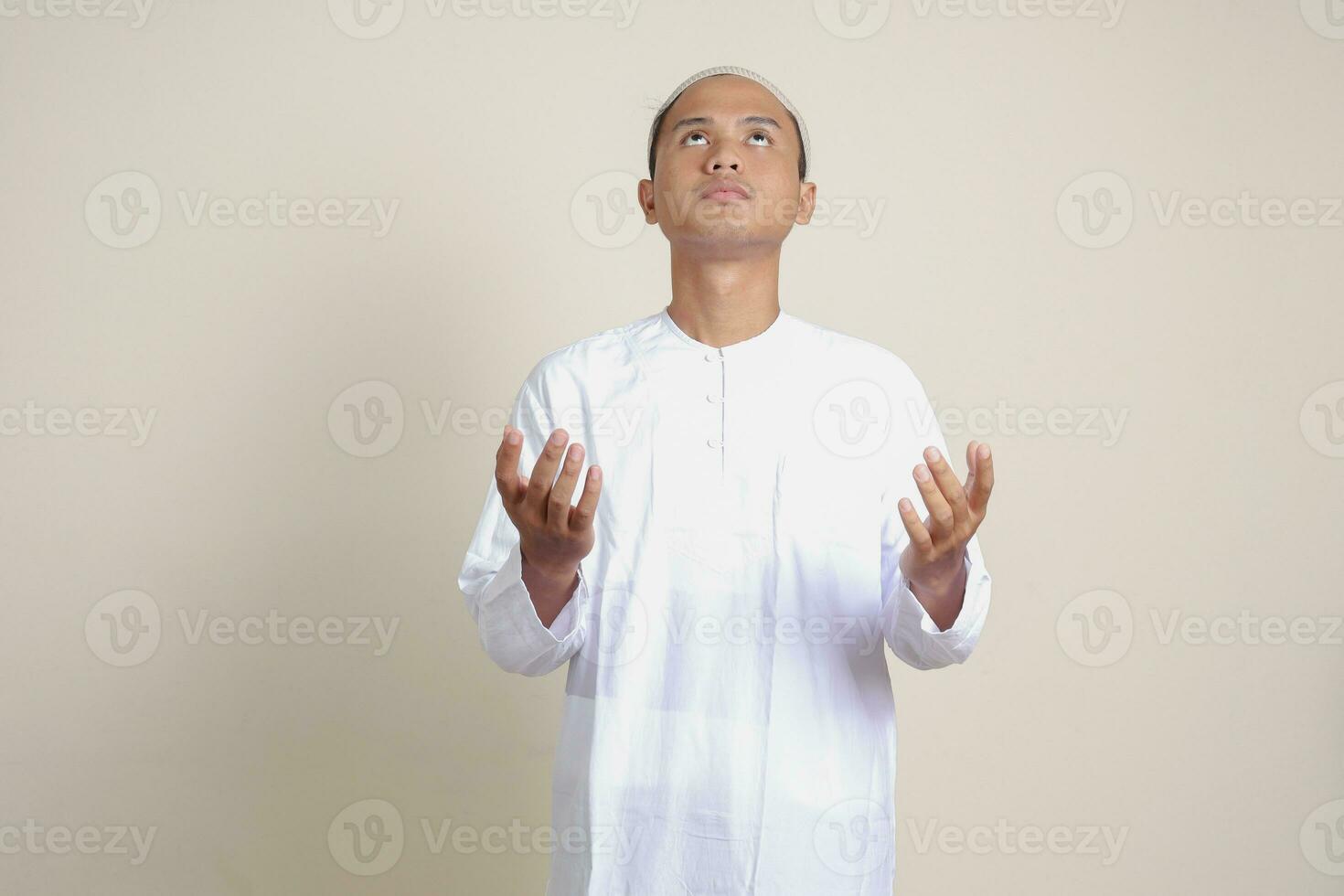 Portrait of attractive Asian muslim man in white shirt with skullcap praying earnestly with his hands raised. Isolated image on gray background photo