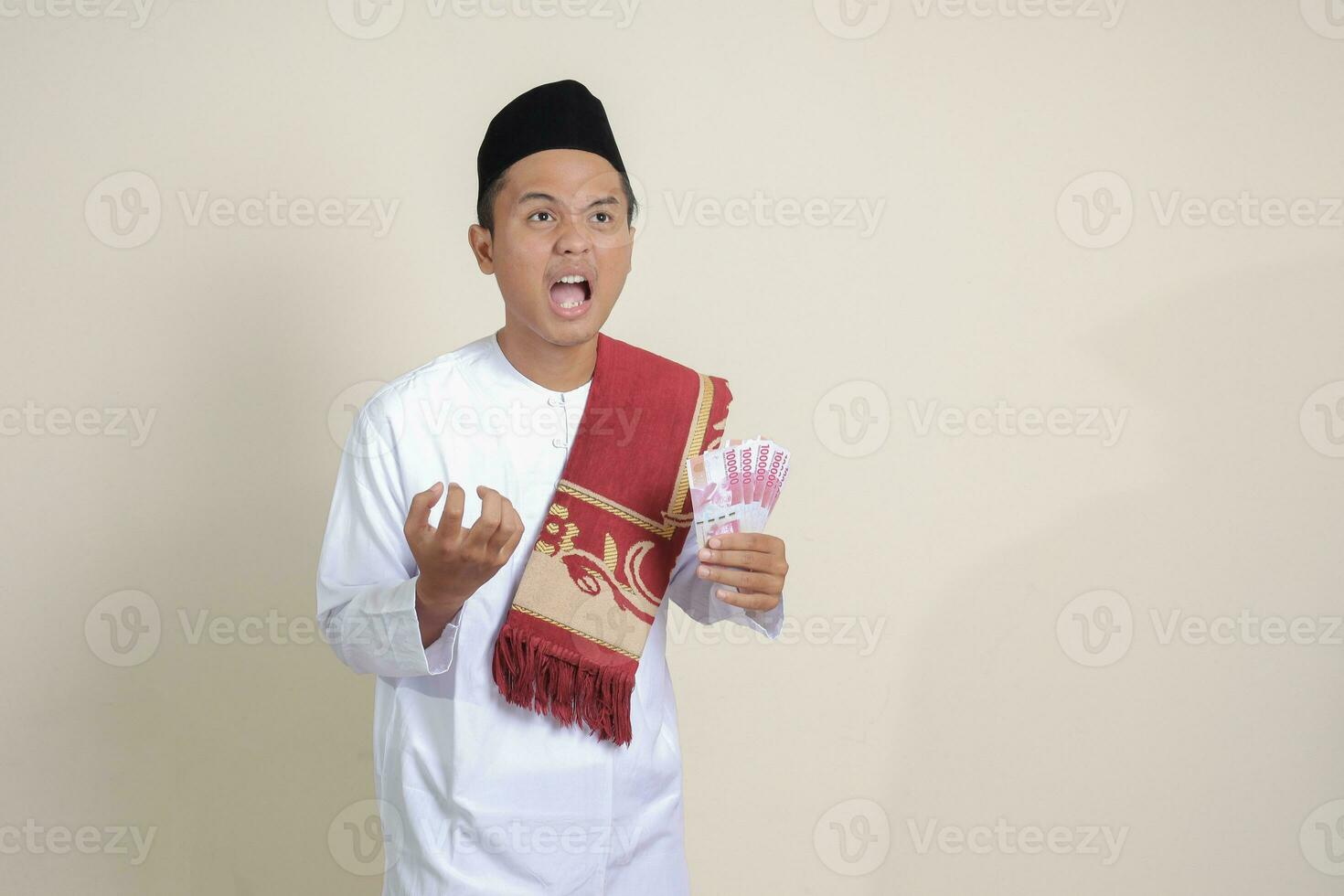 Portrait of attractive Asian muslim man in white shirt making angry gesture showing one hundred thousand rupiah. Financial and savings concept. Isolated image on gray background photo