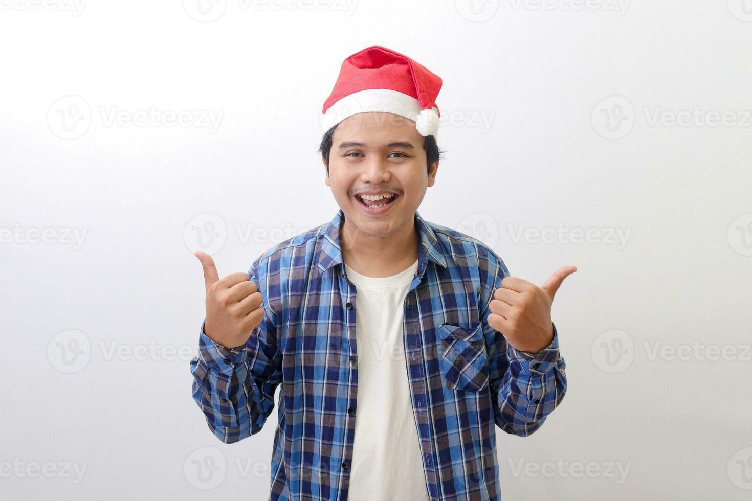 Portrait of excited Asian man in blue plaid shirt wearing santa hat showing thumbs up hand gesture. Isolated image on white background photo