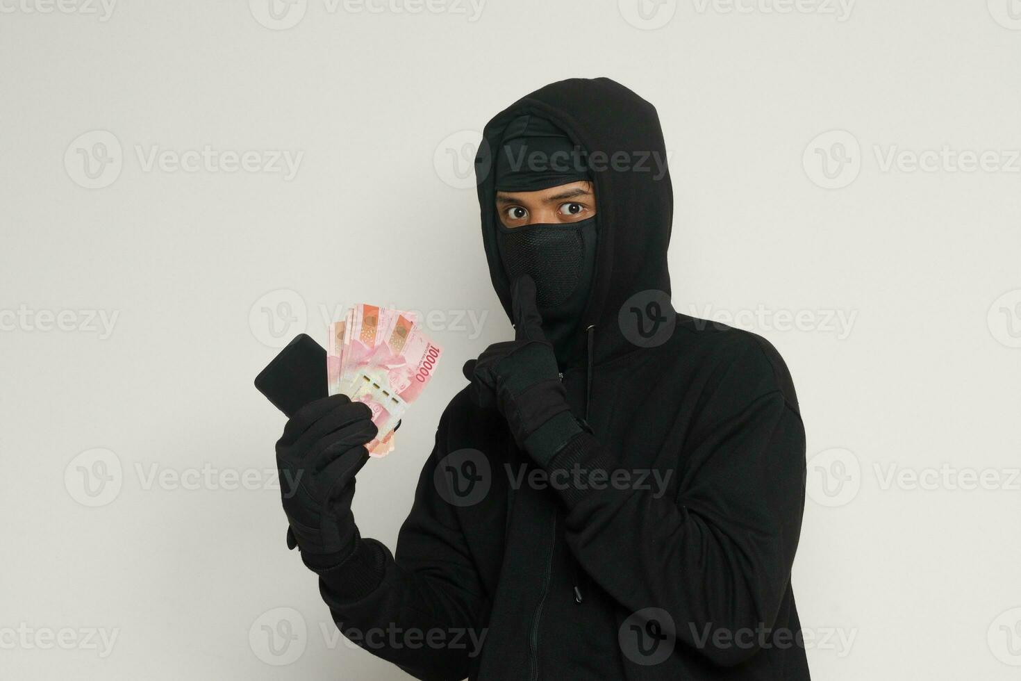 Portrait of mysterious man wearing black hoodie and mask stealing one hundred thousand rupiah successfully from victim. Isolated image on gray background photo