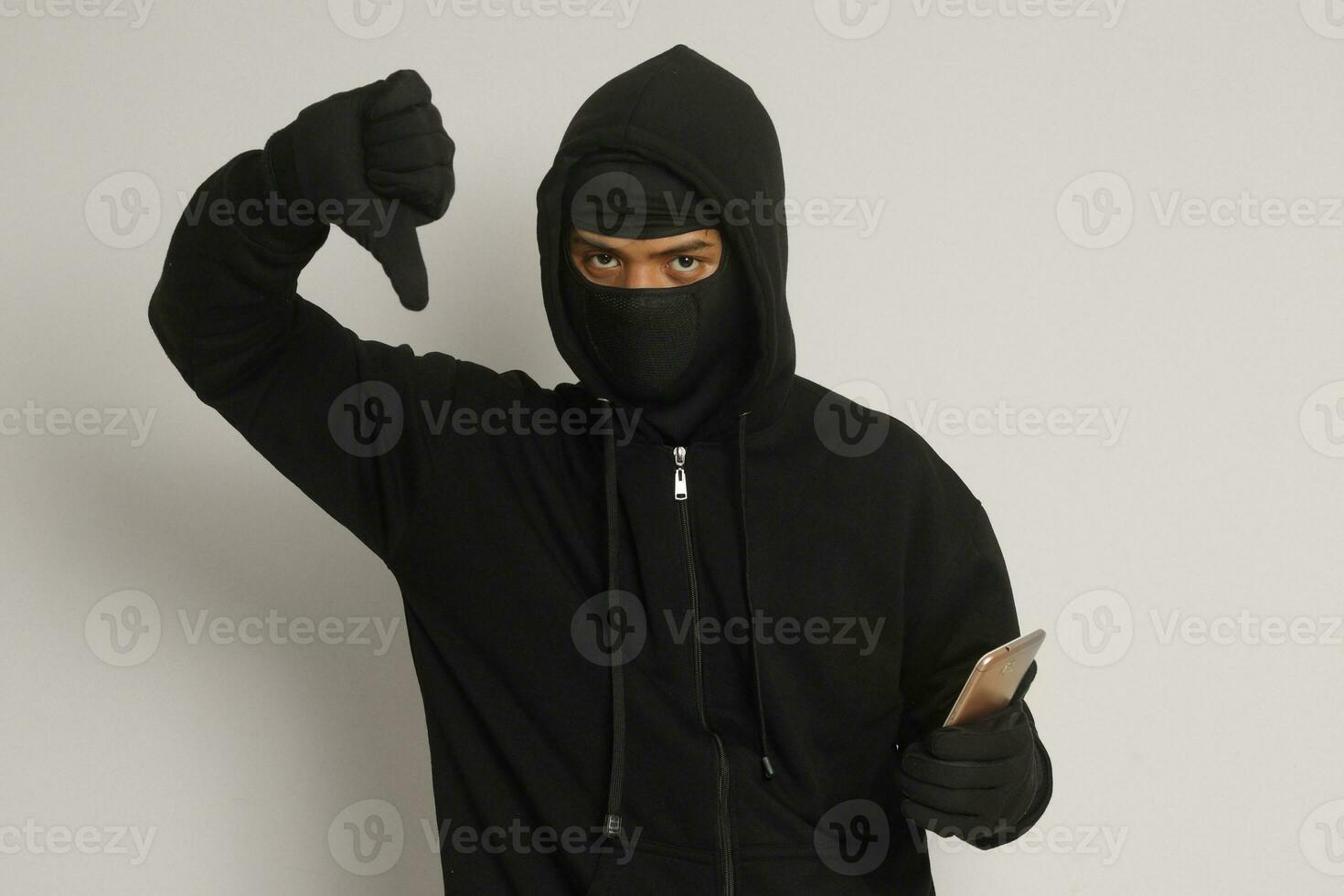 Portrait of mysterious man wearing black hoodie and mask doing hacking activity on mobile phone, hacker holding a smartphone. Isolated image on gray background photo